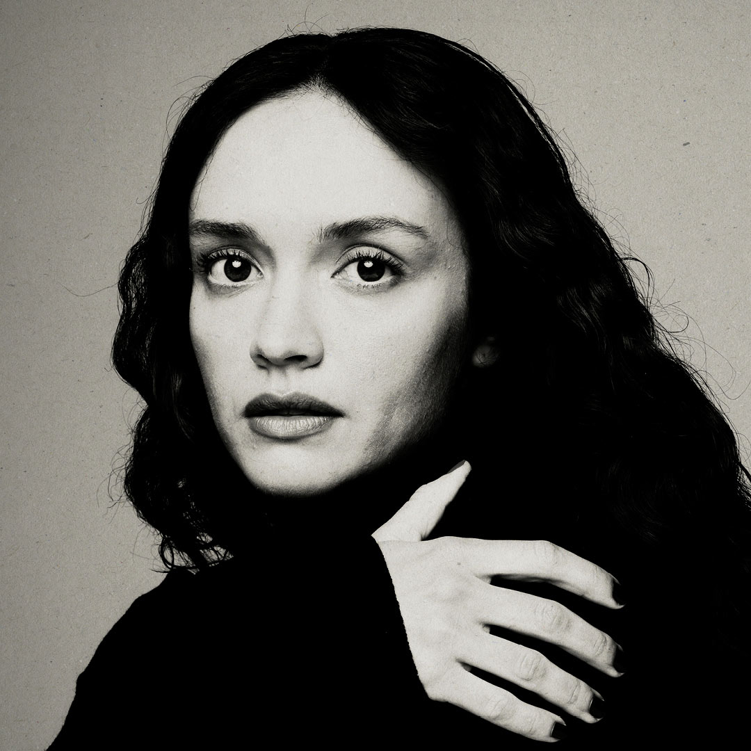 A person with long dark hair poses against a neutral background, looking directly at the camera.