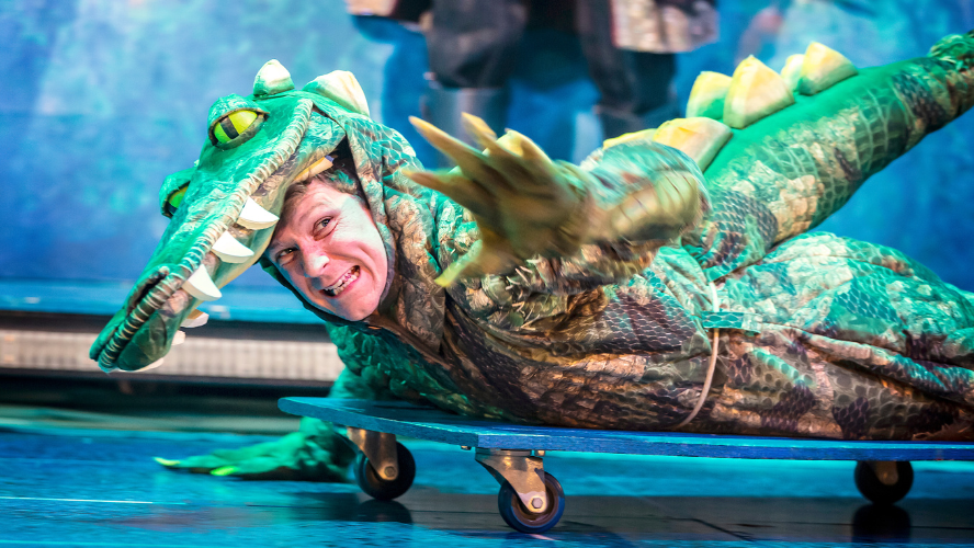 A man in a crocodile costume lies down on a skateboard.